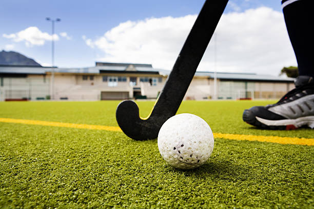 Hockey player, stick and ball on artificial turf. Pavilion and floodlight in the background.