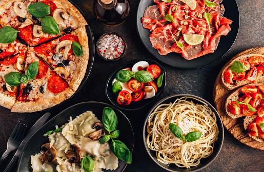 Full table of italian meals on plates Pizza, pasta, ravioli, carpaccio. caprese salad and tomato bruschetta on black background. Top view